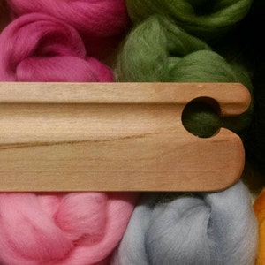 Puede incluir: Una aguja de tejer de madera con un gancho en un extremo. La aguja est&aacute; apoyada sobre una pila de hilo de lana de colores en tonos rosa, verde, azul y amarillo.
