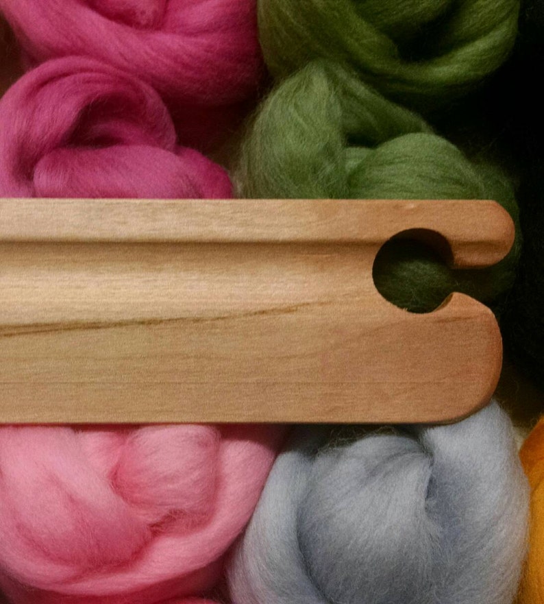 Puede incluir: Una aguja de tejer de madera con un gancho en un extremo. La aguja est&aacute; apoyada sobre una pila de hilo de lana de colores en tonos rosa, verde, azul y amarillo.