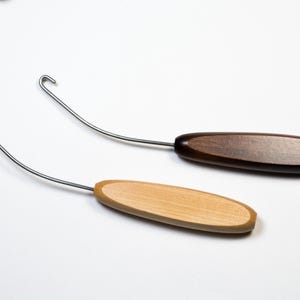 May include: Two wooden handle tools with curved metal hooks. One handle is dark brown, the other is light brown. The tools are likely used for crafts or sewing, and are set against a white background.
