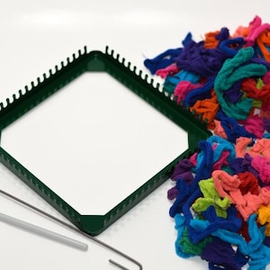 May include: A green square weaving loom with a hook and a pick. Colorful fabric strips in shades of blue, purple, orange, and pink are arranged next to the loom. Craft supplies for weaving.