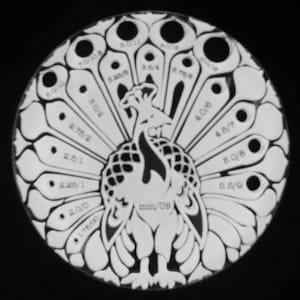 May include: A white peacock design with a black silhouette of the bird. The peacock's tail feathers are numbered with different sizes in inches and fractions of an inch. The design is on a round, white background.