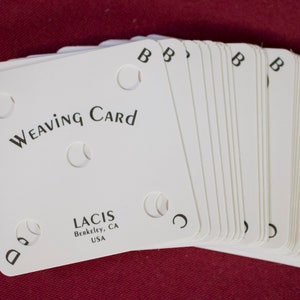 Puede incluir: Un mont&oacute;n de tarjetas de tejido blancas con esquinas redondeadas, con el texto "Weaving Card" y la marca "LACIS". Cada tarjeta tiene cuatro recortes circulares y est&aacute; marcada con las letras A, B, C y D. Las tarjetas est&aacute;n sobre una superficie roja.