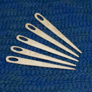 Puede incluir: Cinco agujas de tejer de madera con una punta puntiaguda y un peque&ntilde;o bucle en la parte superior. Las agujas est&aacute;n dispuestas en forma de abanico sobre una tela de punto azul.