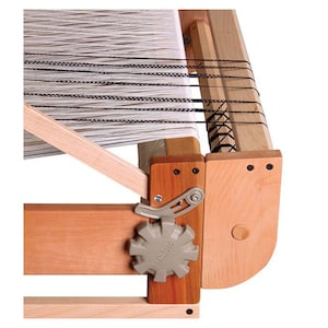 May include: Close-up of a wooden loom with a gray tension knob labeled "Ashford". The loom is set up with white warp threads and black weft threads.