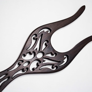 May include: A dark brown wooden hair pick with a decorative, intricate cut-out design. The pick has two prongs at the top and a long, narrow handle.