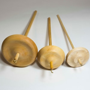 May include: Three wooden spinning wheels of different sizes. The wheels are made of light wood and have a smooth, rounded surface. The spindles are made of a darker wood and have a small hook at the end.