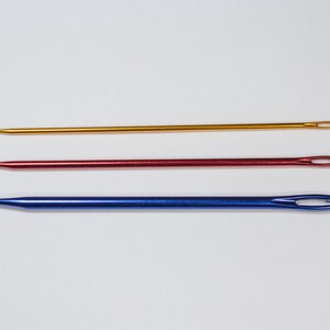 May include: Three crochet hooks in gold, red, and blue. Each hook has a hook on one end and an eyelet on the other. The hooks are arranged horizontally against a white background.
