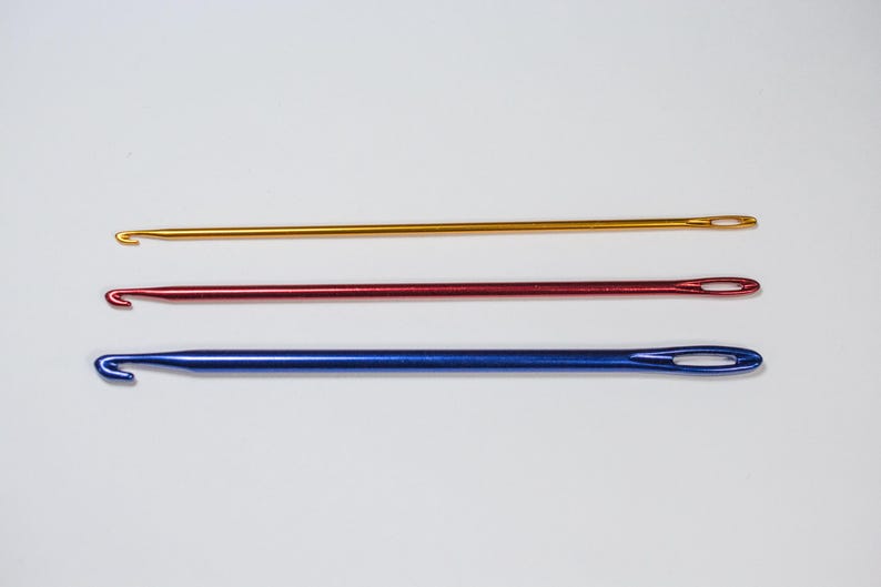 May include: Three crochet hooks in gold, red, and blue. Each hook has a hook on one end and an eyelet on the other. The hooks are arranged horizontally against a white background.
