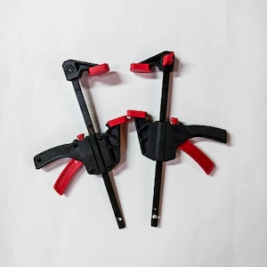May include: Two black and red bar clamps are displayed against a white background. The clamps have a trigger handle and a red clamp pad. The clamps are used for woodworking and construction projects.