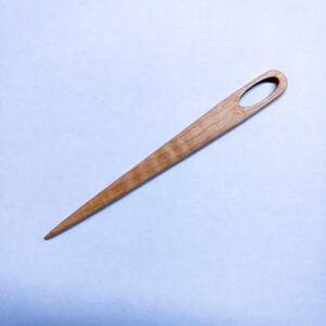 Puede incluir: Un enhebrador de agujas de madera con una punta puntiaguda y un agujero ovalado en la parte superior. El enhebrador est&aacute; hecho de madera marr&oacute;n claro con un patr&oacute;n de veta natural.