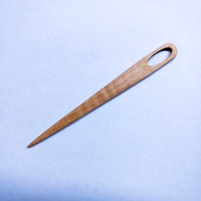 Puede incluir: Un enhebrador de agujas de madera con una punta puntiaguda y un agujero ovalado en la parte superior. El enhebrador est&aacute; hecho de madera marr&oacute;n claro con un patr&oacute;n de veta natural.