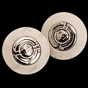 May include: A pair of white ceramic earrings with silver geometric designs. The earrings are round and have a raised design in the center.