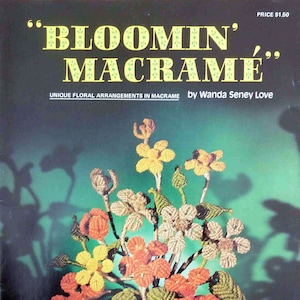 May include: A book titled "Bloomin' Macrame" by Wanda Seney Love, featuring a colorful macrame flower arrangement with orange, yellow, and brown yarn. The book cover is green with a white title and subtitle.