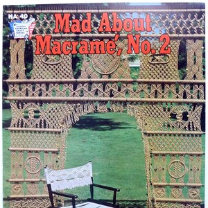 May include: A vintage book cover with the title "Mad About Macrame, No. 2" in red text. The cover features a macrame archway made of natural colored cord, with a white macrame chair and stool in front of it. The background is a green lawn.
