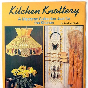 May include: A book cover with the title "Kitchen Knottery: A Macrame Collection Just for the Kitchen" by Pauline Finch. The cover features two macrame projects: a yellow lampshade and a white utensil holder with orange tassels. The book cover is orange with a plaid border.