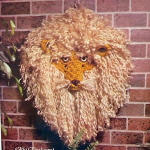 May include: A crocheted lion head wall hanging with a light brown mane and a yellow face. The lion's eyes are black and the nose is brown. The text "Elroy (The Lion)" is printed below the lion's head.