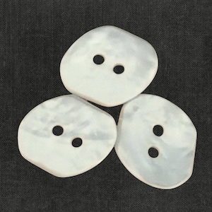 May include: Three off-white, irregular-shaped buttons with two holes each, arranged on a dark gray textured background. The buttons have a slightly pearlescent sheen, suggesting a natural material.