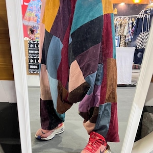 May include: A person wearing a pair of colourful patchwork trousers with a blue shirt and red trainers. The trousers are made of different fabrics and colours, including brown, red, blue, green, and purple.