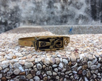 May include: A gold-toned bracelet with a black and gold ankh symbol charm. The bracelet is on a textured surface.