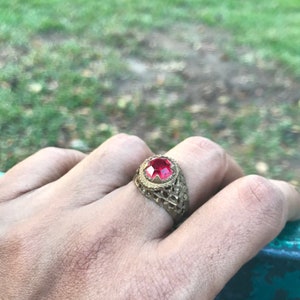 May include: A gold-toned ring featuring a large, round, red gemstone. The ring has an intricate, openwork pattern around the gem and band. The ring is on a hand, with a blurred green backdrop.