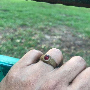 Può includere: Un anello dorato con una gemma rossa rotonda al centro. L'anello è indossato su un dito, con erba verde e una barra di metallo verde sullo sfondo.