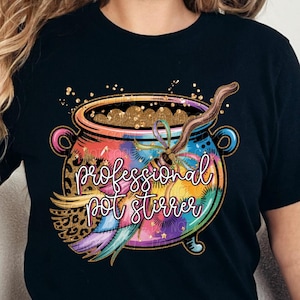 May include: Black t-shirt featuring a colorful cauldron design with the text "professional pot stirrer." The cauldron is filled with gold glitter and a wooden stirring stick. The design includes rainbow colors and a leopard print.