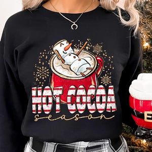 Op de afbeelding: Een zwart sweatshirt met lange mouwen met een afbeelding van een rode mok gevuld met warme chocolademelk en een marshmallow-sneeuwpop. De tekst luidt "HOT COCOA season" in rood-wit gestreepte letters. Een rode mok in de vorm van een kerstmanpak is ook zichtbaar.