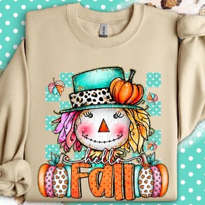 May include: Beige sweatshirt featuring a whimsical scarecrow design with a turquoise hat, orange pumpkin, and the text "hello Fall." The scarecrow has colorful hair and a smiling face. The design includes butterflies and pumpkins.
