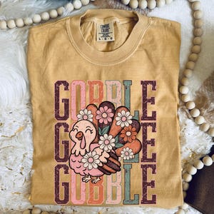 May include: Mustard yellow Comfort Colors t-shirt with a graphic of a cartoon turkey with a floral tail and the word "GOBBLE" in a vertical stack. The turkey is pink and the flowers are pink, white, and orange.