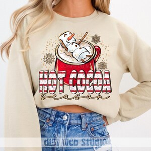 Op de afbeelding: Een beige sweater met ronde hals en een afbeelding van een rode mok gevuld met warme chocolademelk en een marshmallow-sneeuwpop. De tekst "HOT COCOA season" staat eronder in rood-wit gestreepte letters met gouden glitteraccenten.