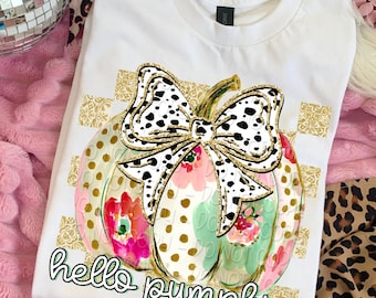 May include: White t-shirt featuring a colorful pumpkin design with a dalmatian print bow and floral accents. The words "hello pumpkin" are written in a script font below the pumpkin. A festive fall-themed graphic tee.