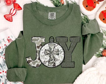 Könnte beinhalten: Ein olivgrünes Langarmshirt mit dem Wort "JOY" in einer dekorativen Schriftart. Das "J" ist Camouflage, das "O" ist eine Weihnachtskugel und das "Y" ist grau. Das Shirt hat den Text "DO NOT COPY" auf der rechten Seite.