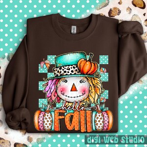 May include: A brown sweatshirt featuring a whimsical scarecrow design with a turquoise hat, orange pumpkin, and the words "hello FALL". The scarecrow has colorful hair and is set against a teal and white checkered background. Two pumpkins flank the text.