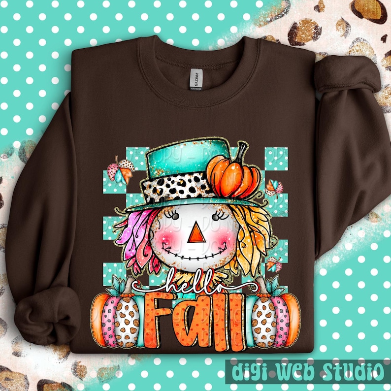 May include: A brown sweatshirt featuring a whimsical scarecrow design with a turquoise hat, orange pumpkin, and the words "hello FALL". The scarecrow has colorful hair and is set against a teal and white checkered background. Two pumpkins flank the text.