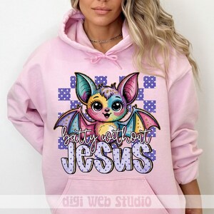 May include: A light pink hooded sweatshirt featuring a colorful cartoon bat with the text "batty without Jesus." The bat has large eyes and multi-colored wings. The background is a checkered pattern of purple and white squares.