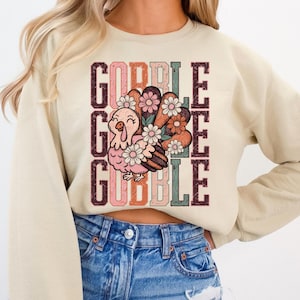 May include: Beige sweatshirt with a colorful graphic of a turkey and the words "GOBBLE GOBBLE" in a vertical layout. The turkey is pink and brown, decorated with flowers. The text is in shades of brown, orange, and pink.