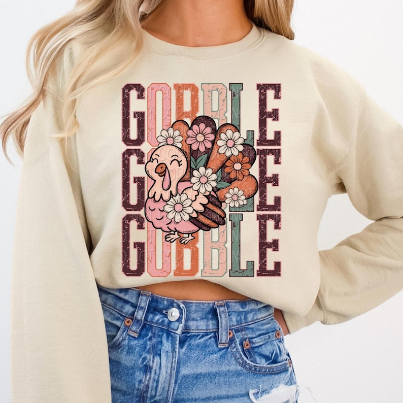 May include: Beige sweatshirt with a colorful graphic of a turkey and the words "GOBBLE GOBBLE" in a vertical layout. The turkey is pink and brown, decorated with flowers. The text is in shades of brown, orange, and pink.