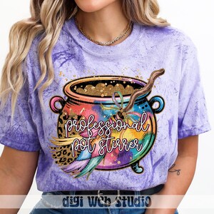 May include: A purple tie-dye t-shirt featuring a colorful cauldron design with the text "professional pot stirrer". The cauldron is filled with gold glitter and has a wooden stirring spoon. The design includes a leopard print and feathers.