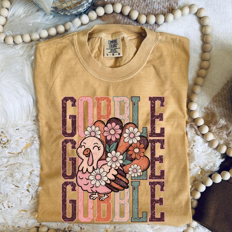 May include: Mustard yellow Comfort Colors t-shirt with a graphic of a cartoon turkey with a floral tail and the word "GOBBLE" in a vertical stack. The turkey is pink and the flowers are pink, white, and orange.
