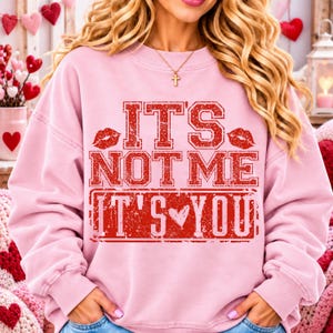 Puede incluir: Una sudadera de cuello redondo en color rosa claro con un estampado gráfico rojo desgastado que dice IT'S NOT ME IT'S YOU, junto a dos estampados de labios rojos y un pequeño icono de corazón. El texto aparece en letras grandes y negritas.