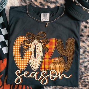 以下が含まれることがあります： 秋をテーマにしたデザインの「COZY season」という文字がプリントされた、濃いグレーのComfort Colors Tシャツ。文字はチェック柄、ヒョウ柄、秋の葉で埋められています。金のネックレスと黒いキャップも写っています。