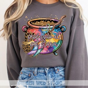 May include: Gray sweatshirt featuring a colorful cauldron design with the text "professional pot stirrer." The cauldron is decorated with a leopard print, feathers, and a wooden stirring stick. Gold glitter accents the design.