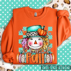 May include: Orange sweatshirt with a colorful scarecrow graphic. The scarecrow wears a teal hat with a leopard print band and holds two pumpkins. The words "hello Fall" are written across the bottom. The design includes butterflies and a checkered background.