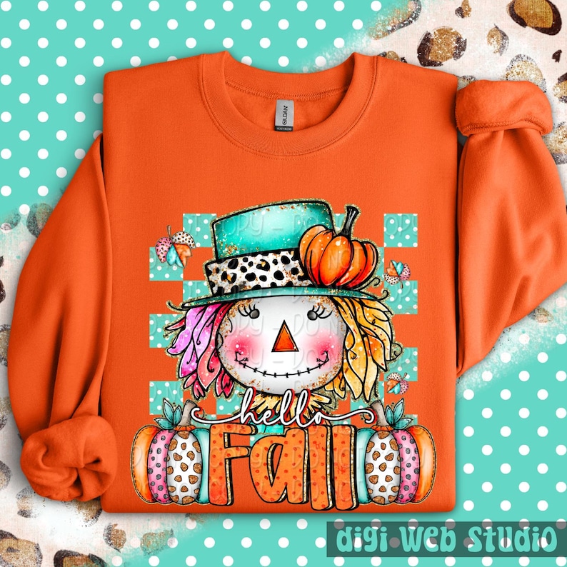 May include: Orange sweatshirt with a colorful scarecrow graphic. The scarecrow wears a teal hat with a leopard print band and holds two pumpkins. The words "hello Fall" are written across the bottom. The design includes butterflies and a checkered background.