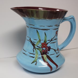 May include: A light blue ceramic pitcher featuring a dark maroon interior rim and a hand-painted floral design. The exterior displays horizontal brown brushstroke bands and a central motif of green leaves with small red and purple flowers.