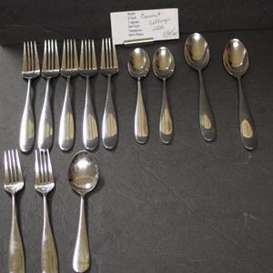 May include: A set of stainless steel flatware, including forks and spoons, arranged on a dark surface. The set includes dinner forks, teaspoons, and tablespoons. A handwritten note is visible in the background.