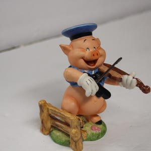 May include: A ceramic figurine of a pig playing a violin. The pig is wearing a blue sailor hat, vest, and white gloves. It stands on a green base with a small wooden fence.