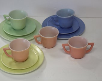 May include: A set of pastel-colored teacups and saucers. The cups are made of glass and have a white interior. The saucers are also made of glass and have a matching color to the cups. The set includes a light green, light blue, and two peach-colored cups and saucers.