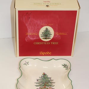 May include: A white ceramic serving dish with a green rim and a green Christmas tree design. The dish is in its original box, which is red with white text that reads "Christmas Tree Spode Since 1938 Celebrating Traditions".