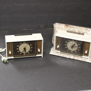 May include: Two vintage electric alarm clocks. The clocks have a white frame with a dark brown clock face and wood-tone side panels. One clock is unwrapped, and the other is in its original packaging.